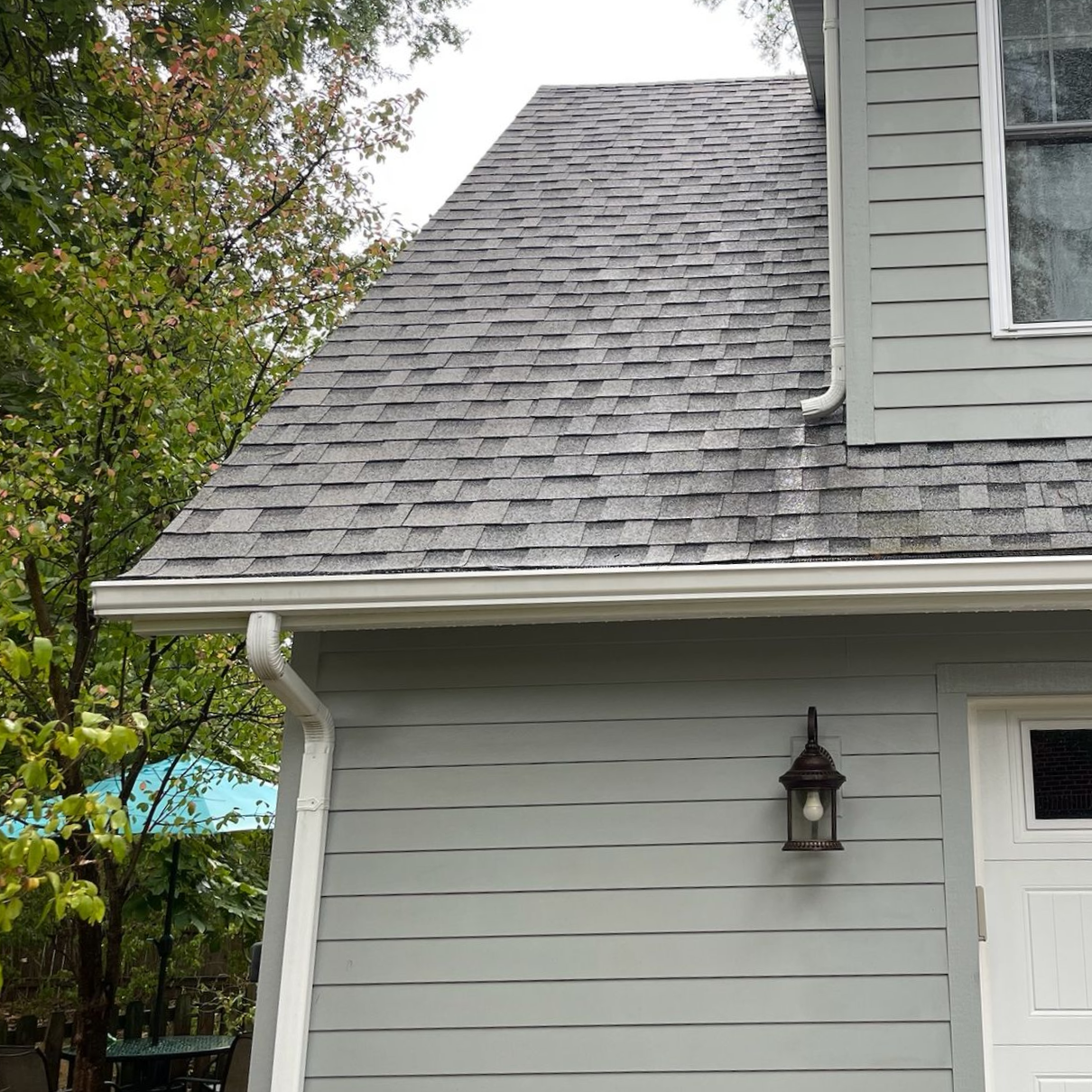 Gray house exterior with shingled roof, white siding, porch light, and trees in the background
