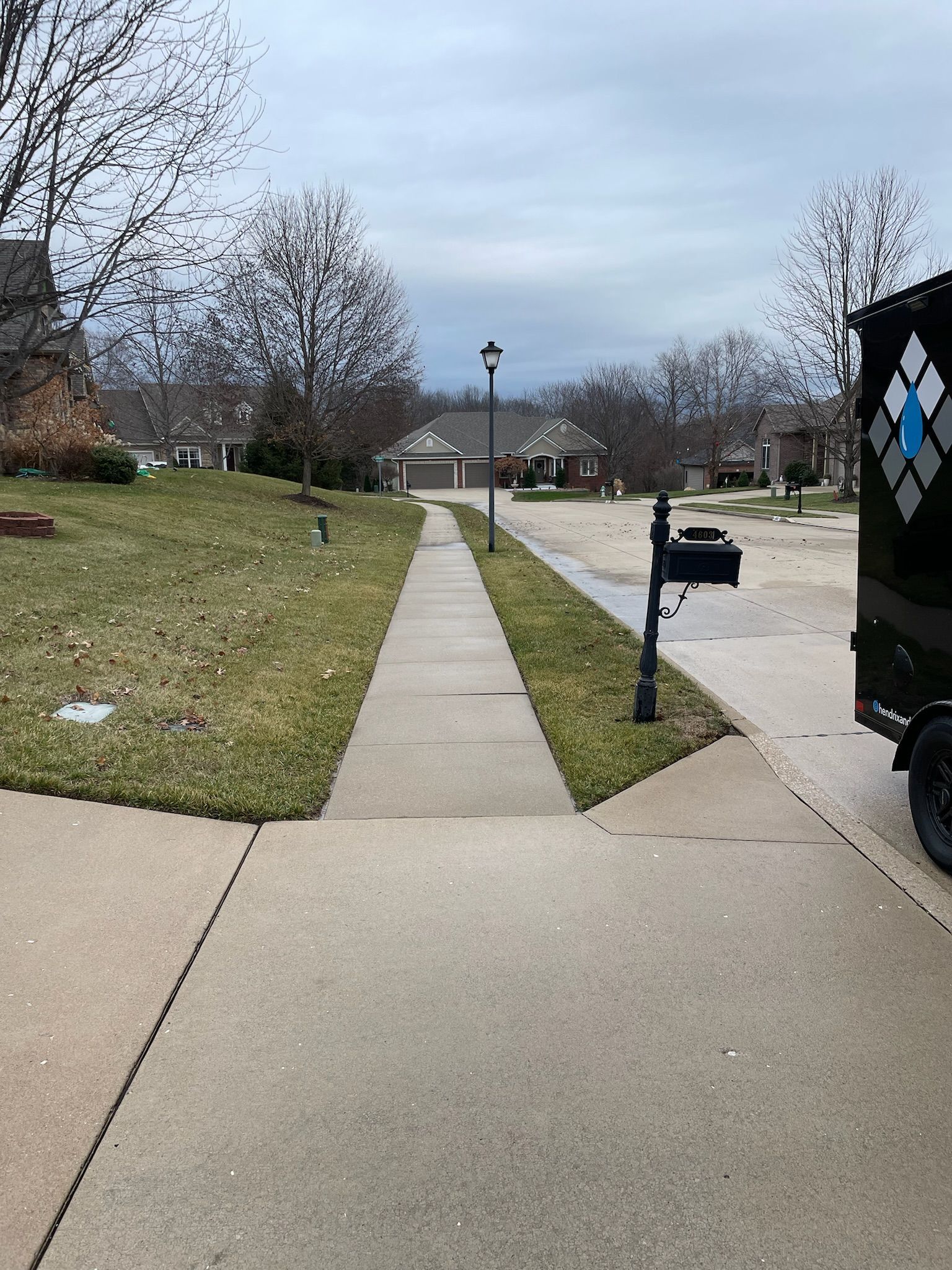 Concrete walkway through a grassy residential area with leafless trees and a mailbox under a cloudy sky.