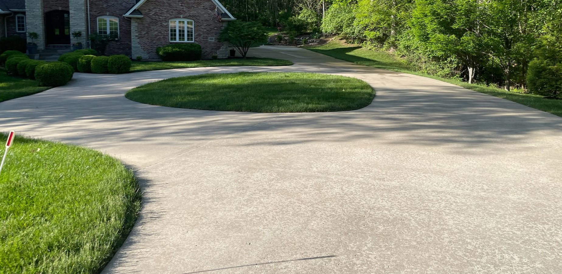 Curved concrete driveway leading to a brick house, bordered by green lawn and trees.