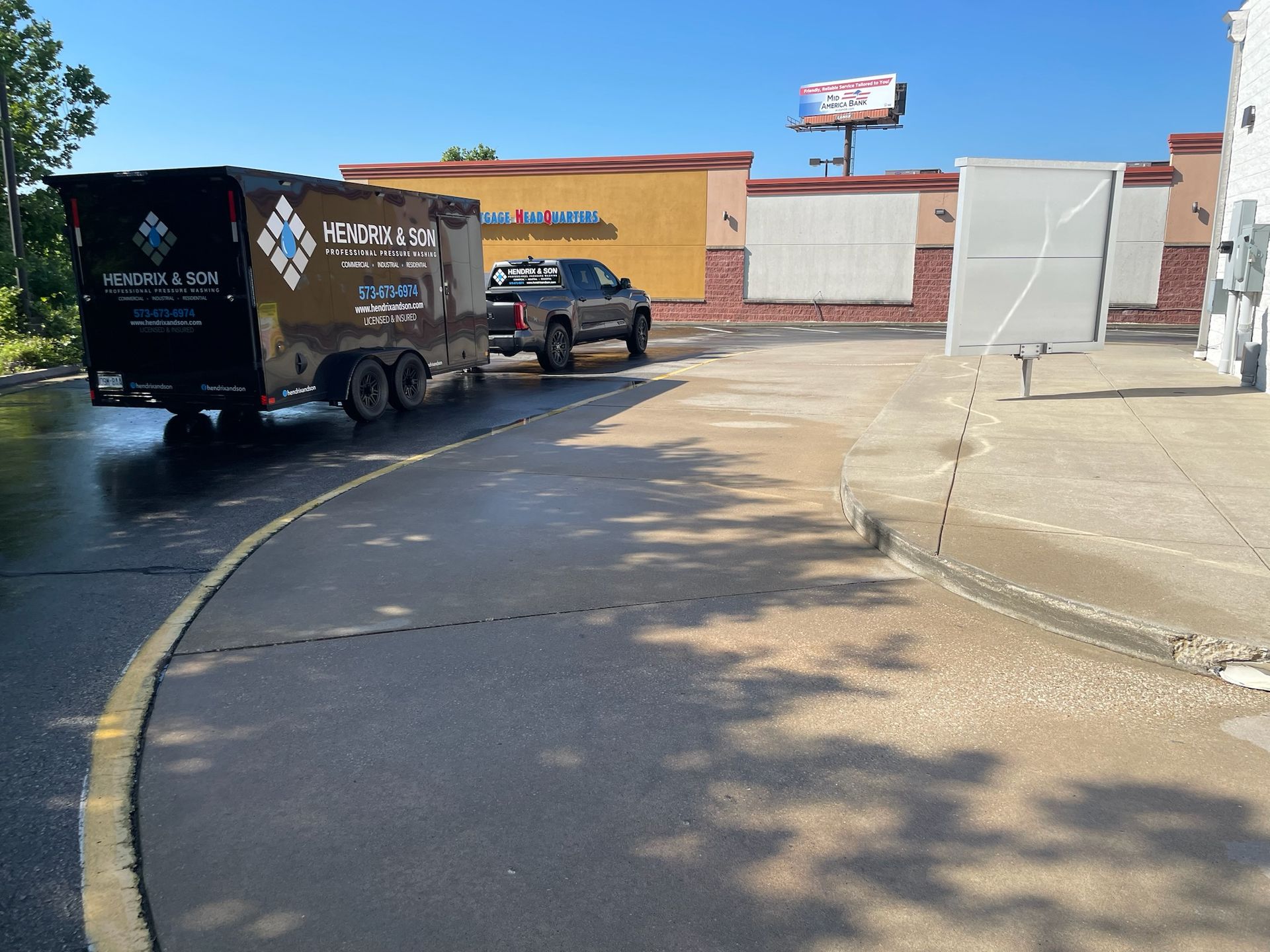 Two trucks parked outside a tan building on a sunlit street with shadows and a billboard overhead