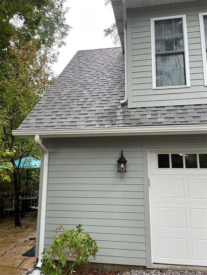 Gray house exterior with white garage door, shingled roof, and wall lantern beside a small shrub