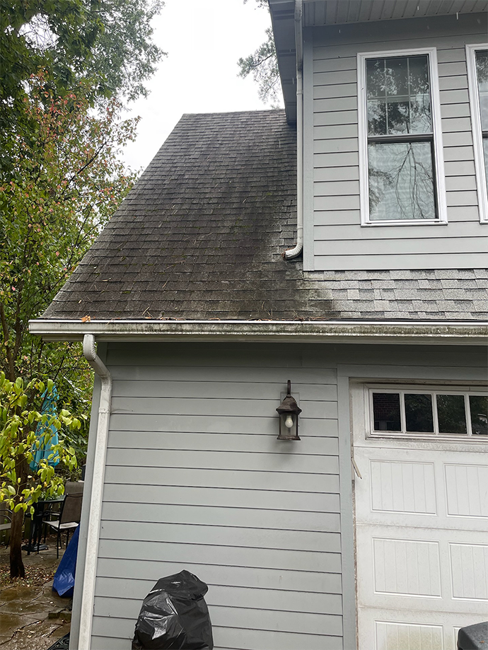 Gray house exterior with steep roof, white garage door, and front window under trees