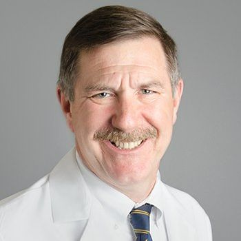 A man with a mustache is wearing a white coat and tie and smiling.