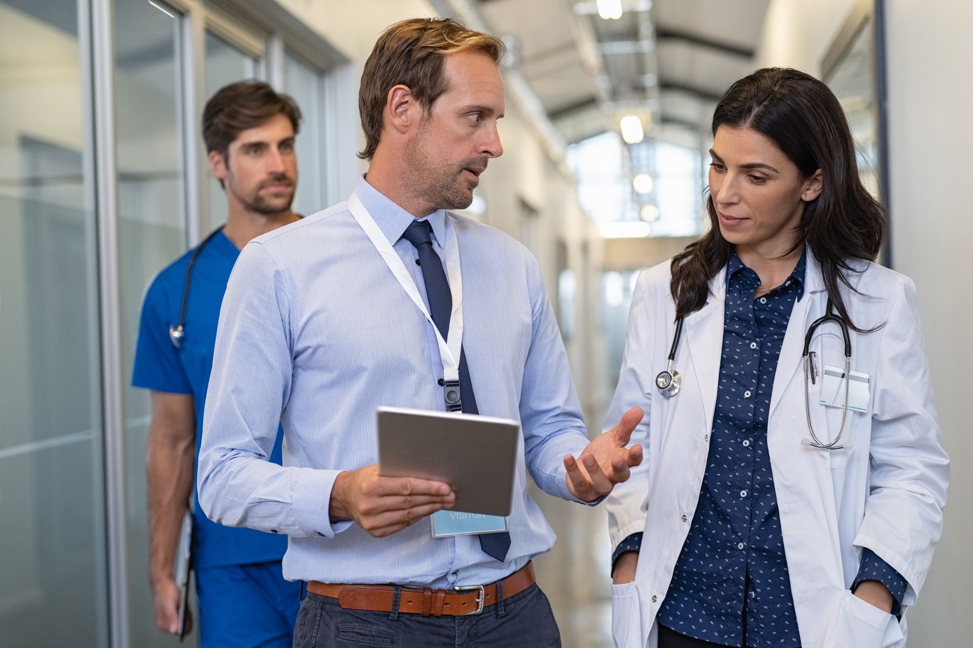 A man is holding a tablet and talking to a woman in a hospital hallway.