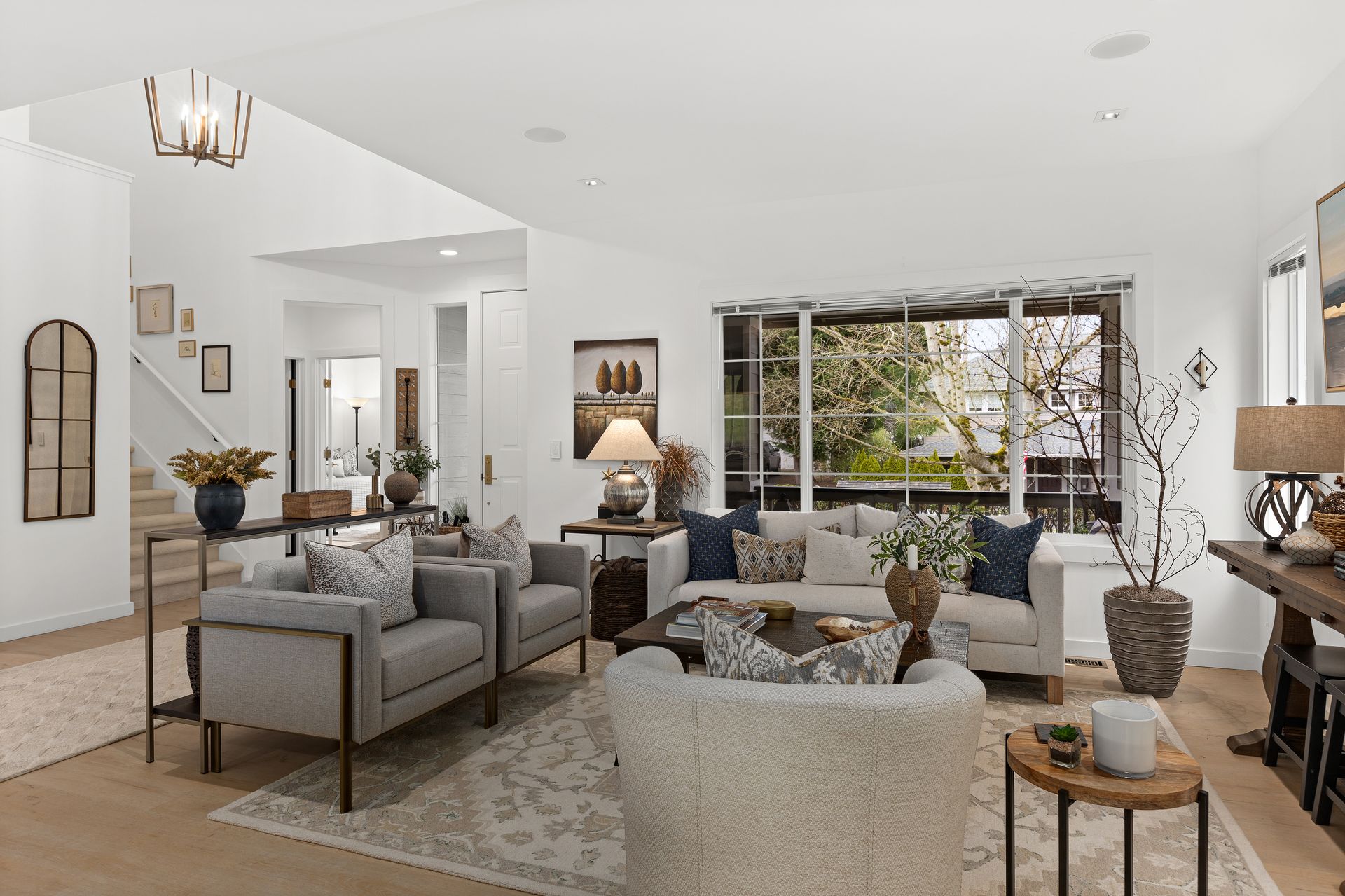 Bright living room with gray sofas, cream chairs, patterned rug, and large window overlooking greenery