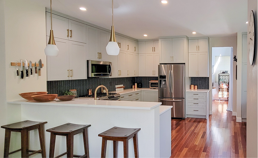Modern white kitchen with island, pendant lights, wood floors, and bar stools; hallway visible in back.