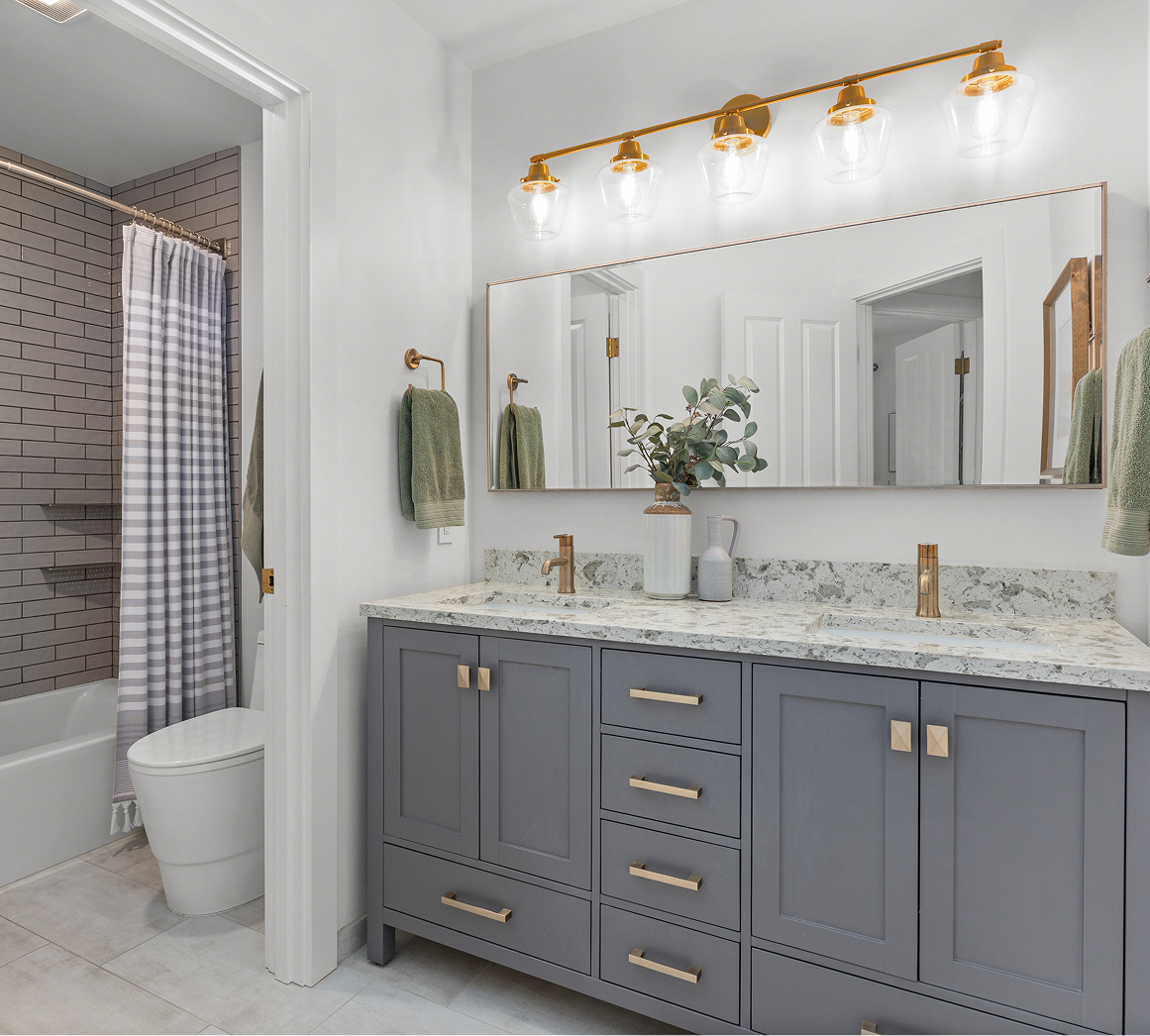 Bright bathroom with gray vanity, marble countertop, gold fixtures, and a tiled shower visible through the doorway.