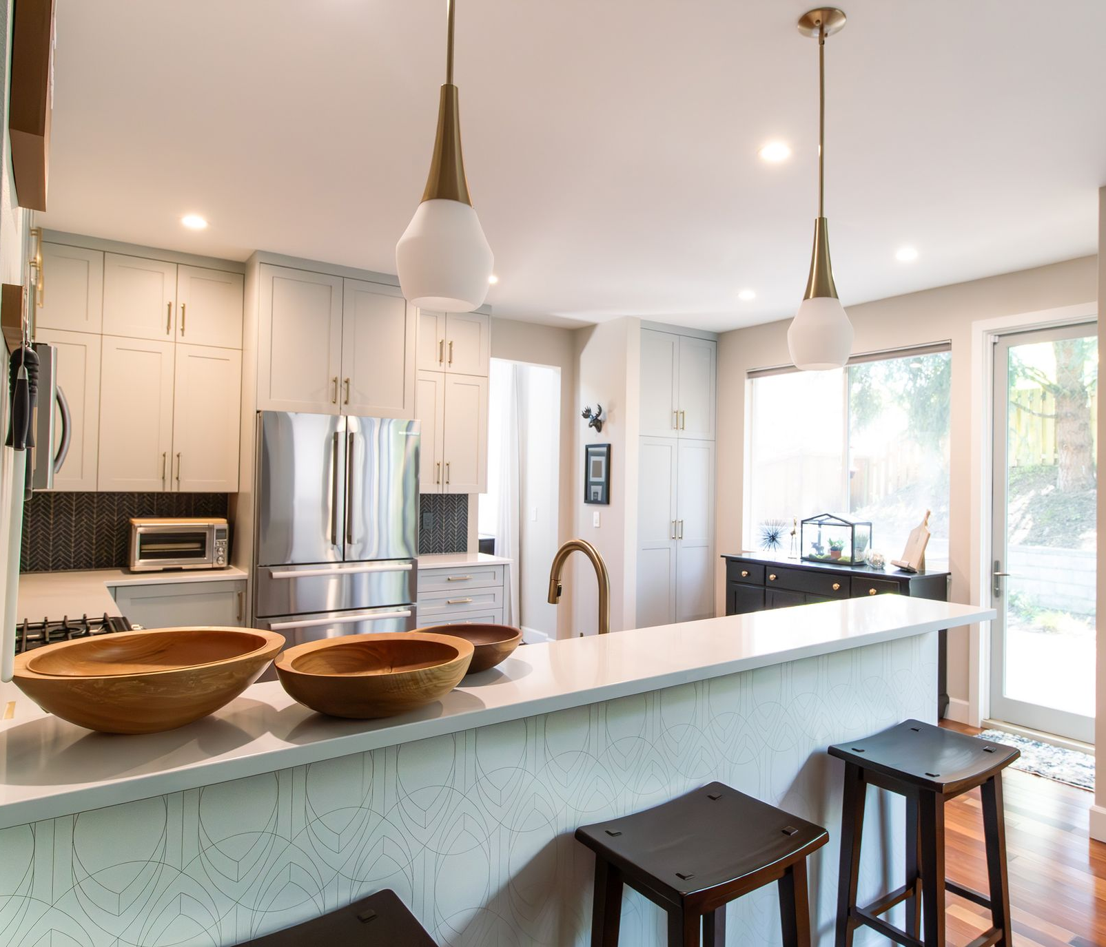 Bright modern kitchen with white cabinets, island stools, pendant lights, and large windows.