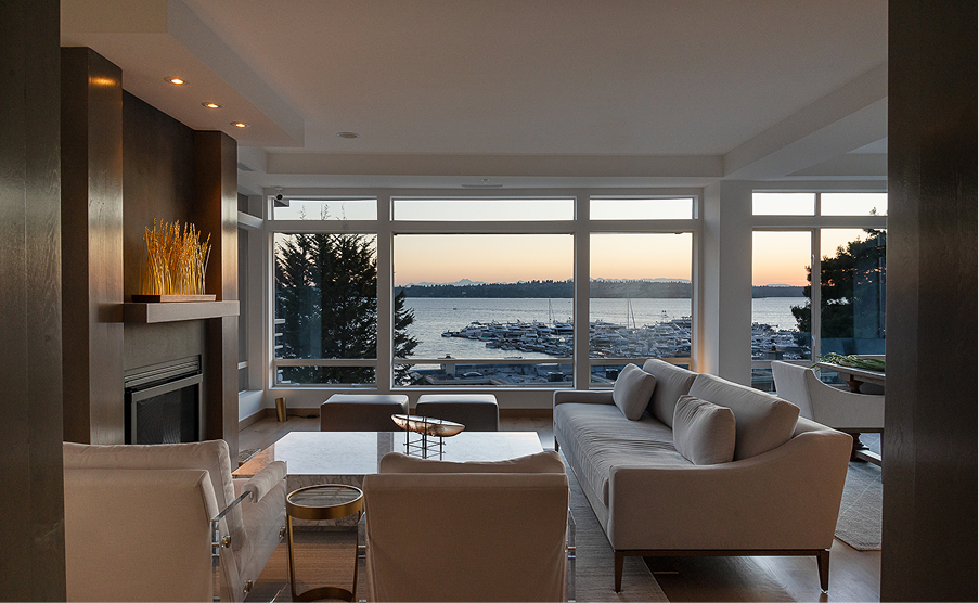 Modern living room with fireplace, sofa, and large windows overlooking a calm waterfront sunset