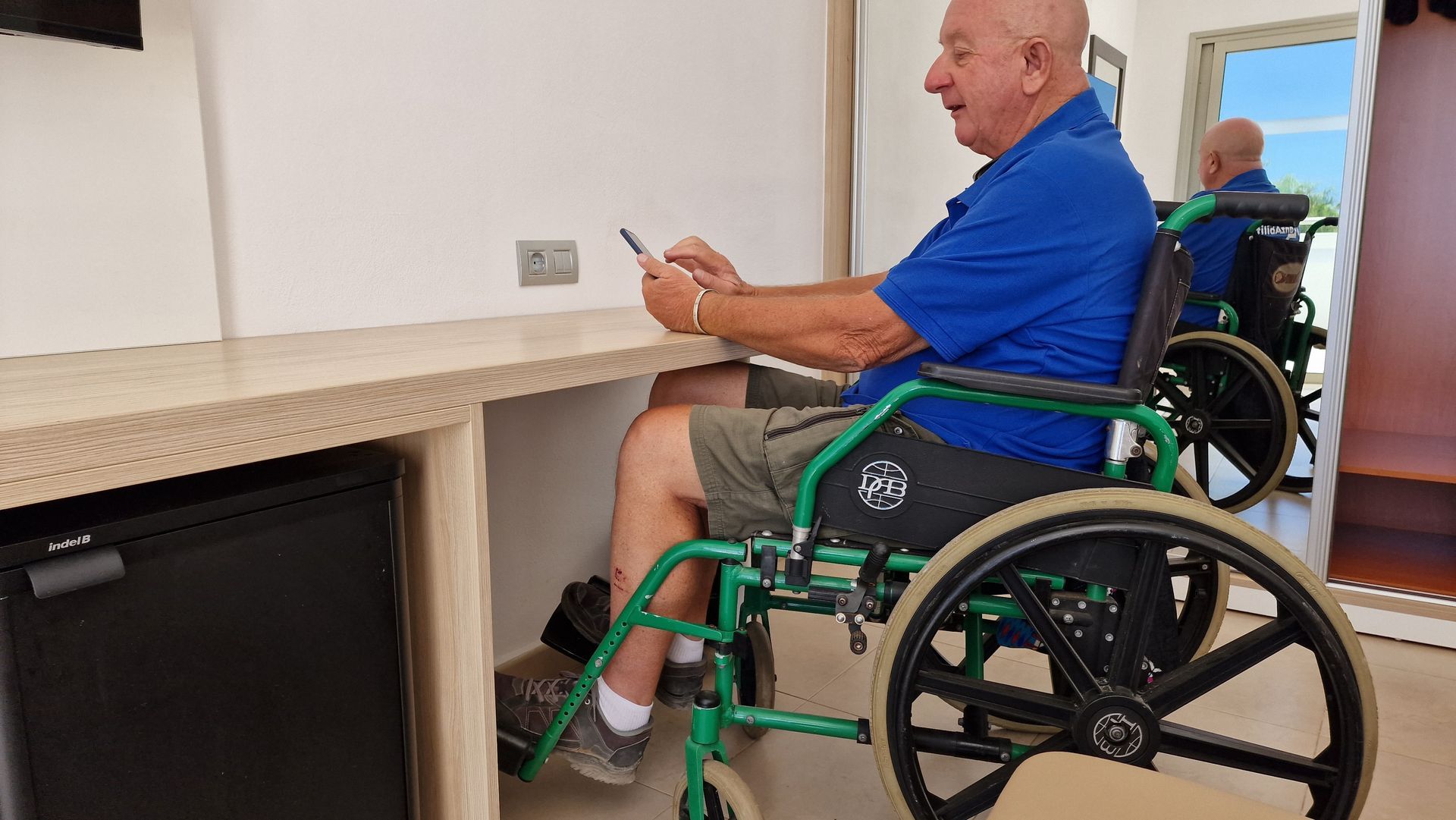 A man in a wheelchair is sitting at a desk using a cell phone.