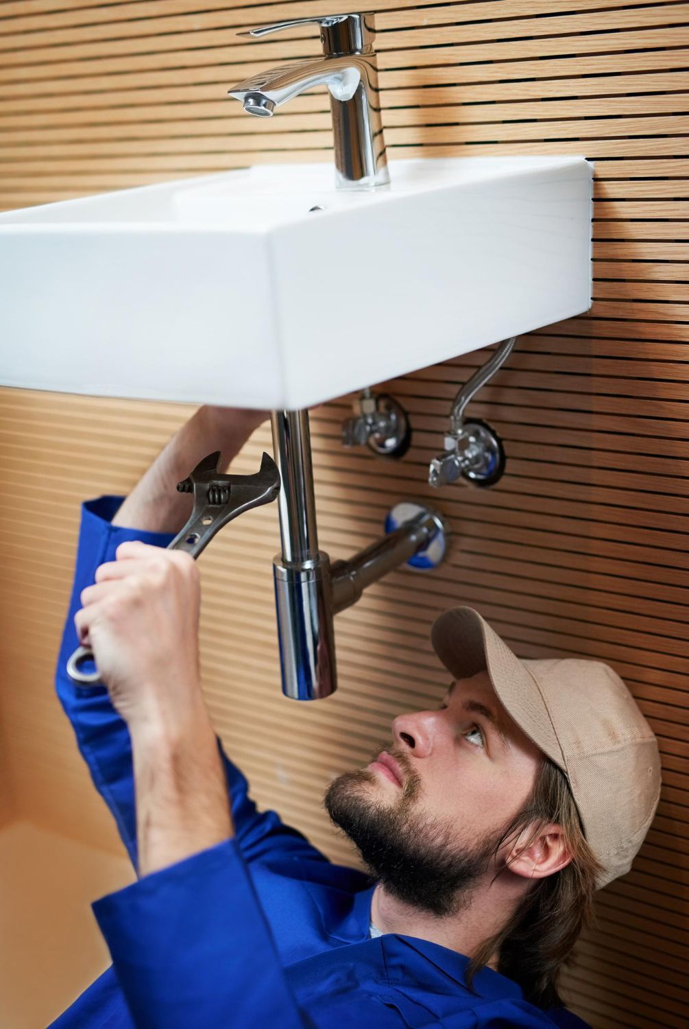 Plumber in blue shirt repairs pipes under a wall-mounted sink in a bathroom.