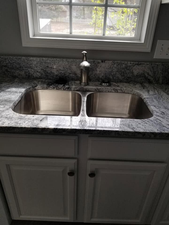 Double stainless steel kitchen sink beneath a window, with gray granite countertop and white cabinets