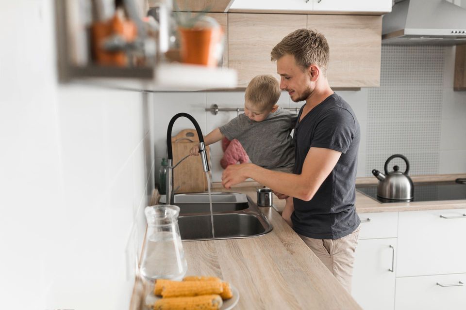 Adult holding a child at a kitchen sink, washing hands in a bright modern kitchen.
