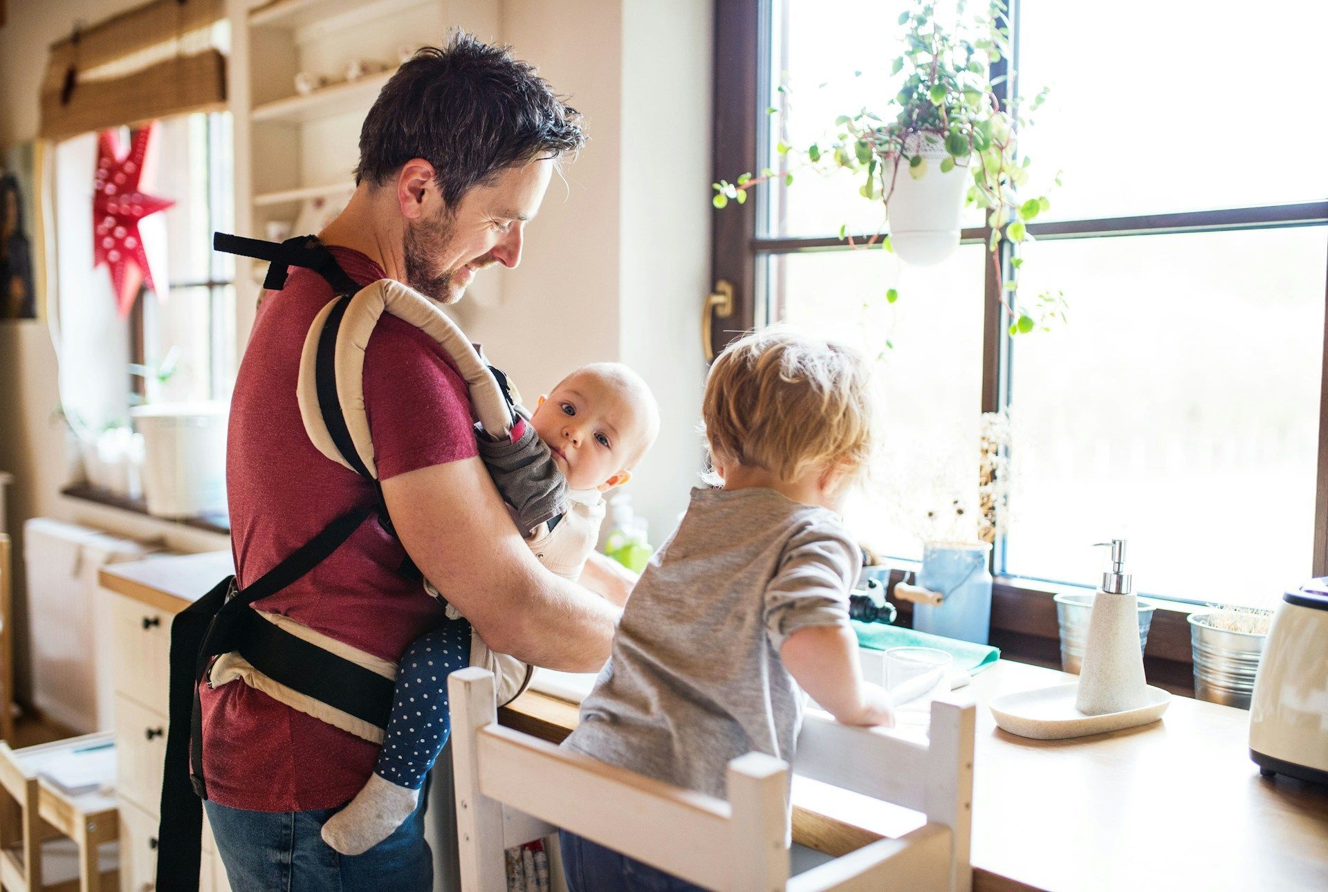 Man in a baby carrier holding two children at a bright kitchen table by a sunlit window