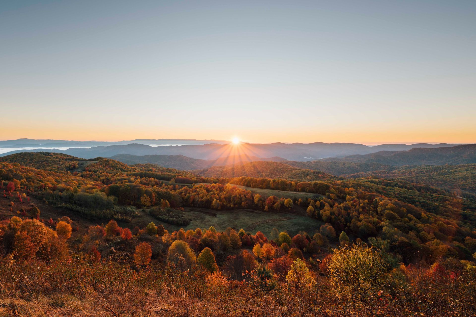 Sunrise over rolling autumn hills with orange foliage and a distant lake under a clear sky