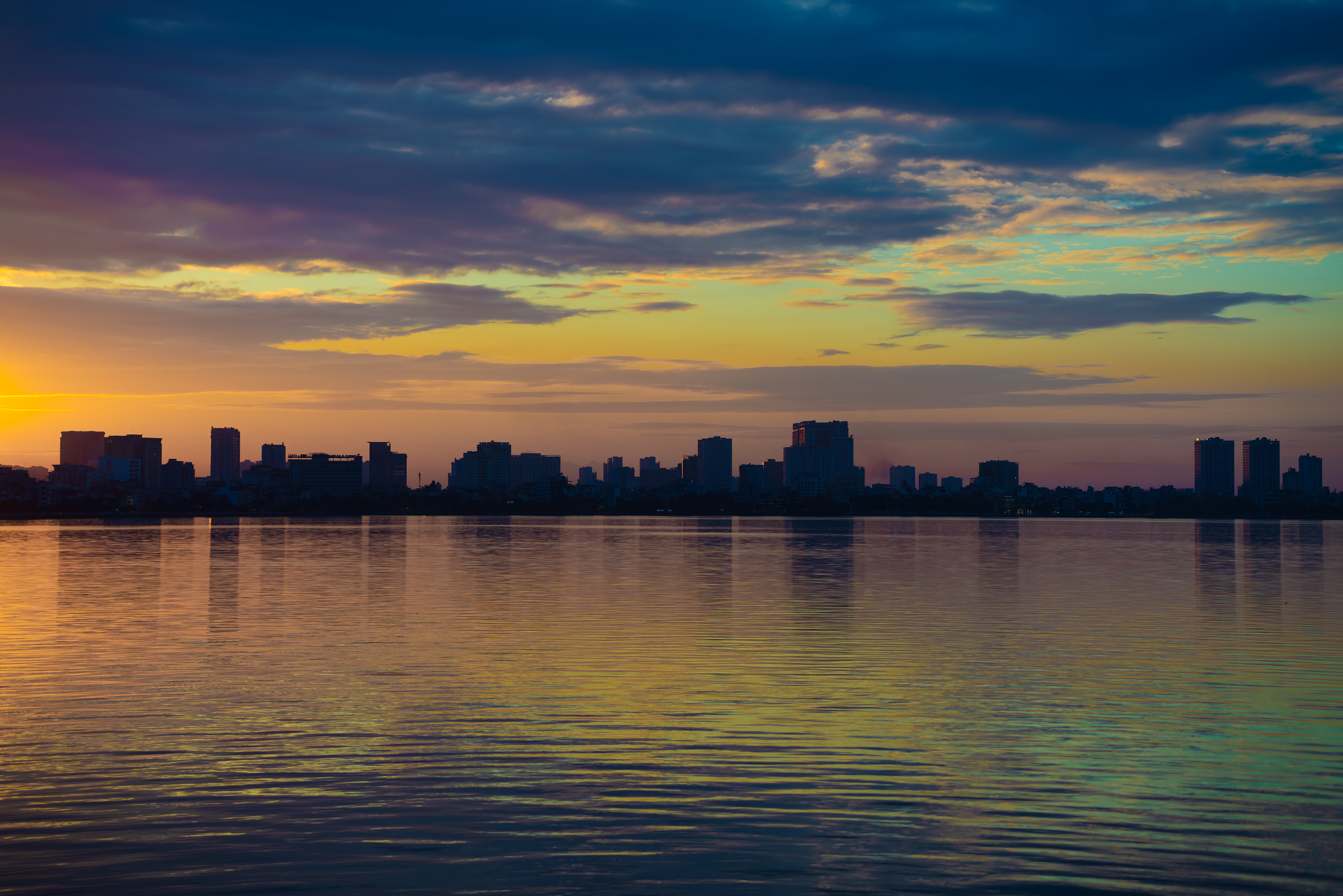 City skyline silhouetted at sunset over calm water, with pink, orange, and purple clouds reflecting below