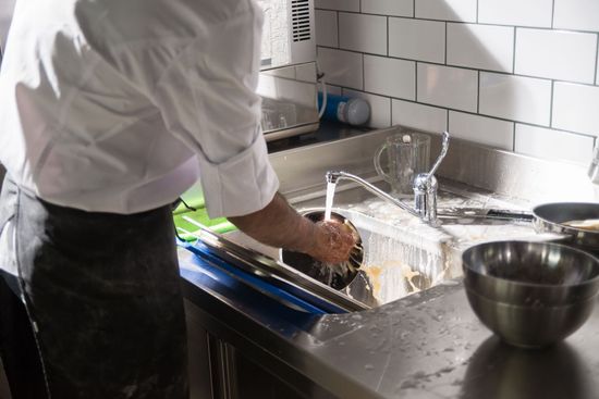 Person washing dishes at a kitchen sink beside a metal bowl and white tile backsplash