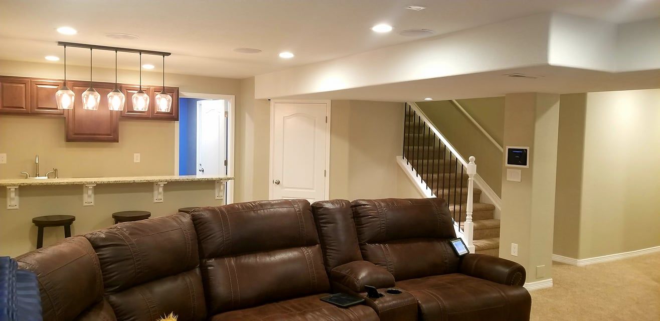 A basement interior with a brown leather sectional, a bar area with wooden cabinets and stools, and stairs in the background.