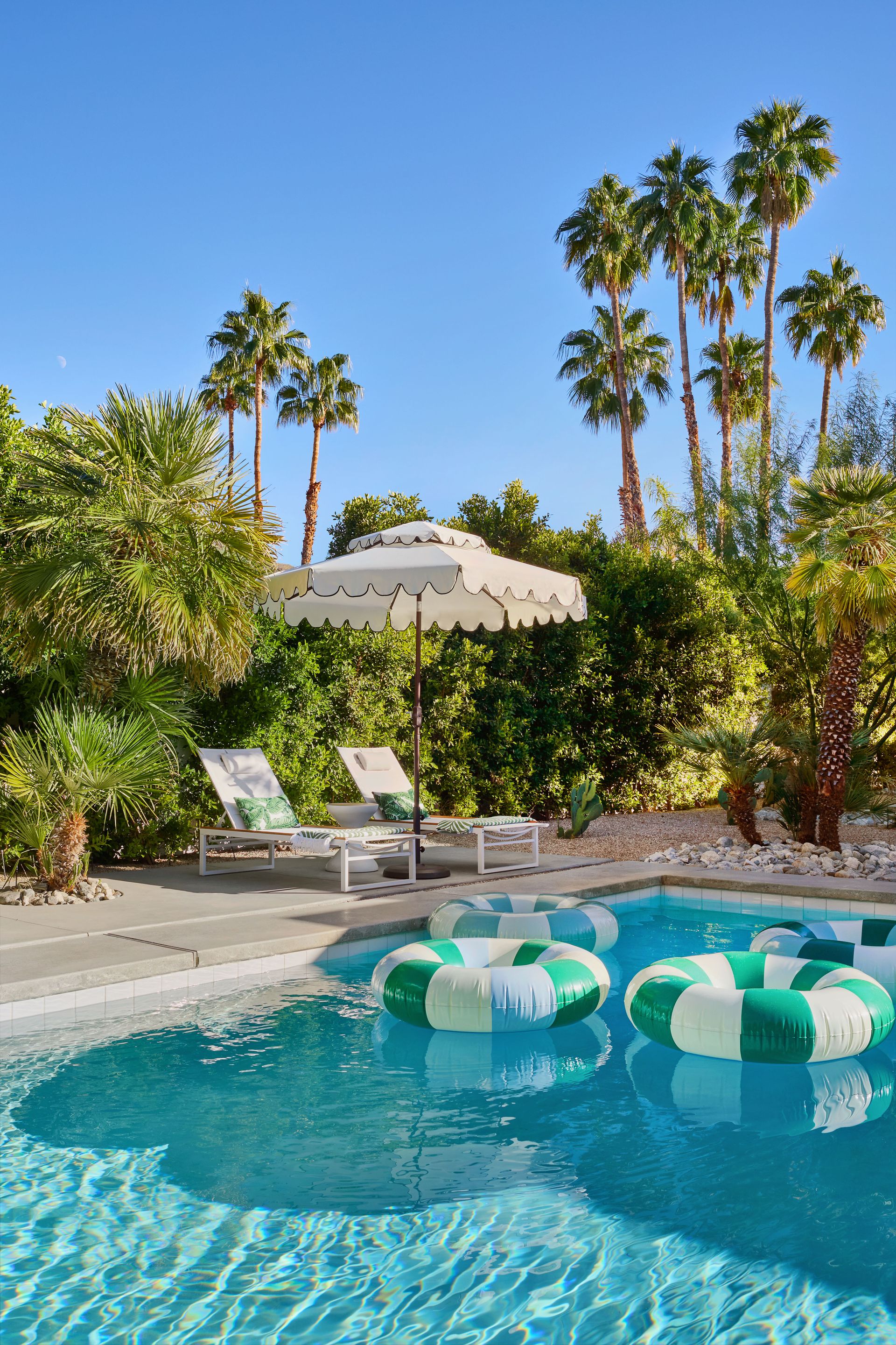 Pool with green and white floats, lounge chairs, umbrella, and palm trees on a sunny day.