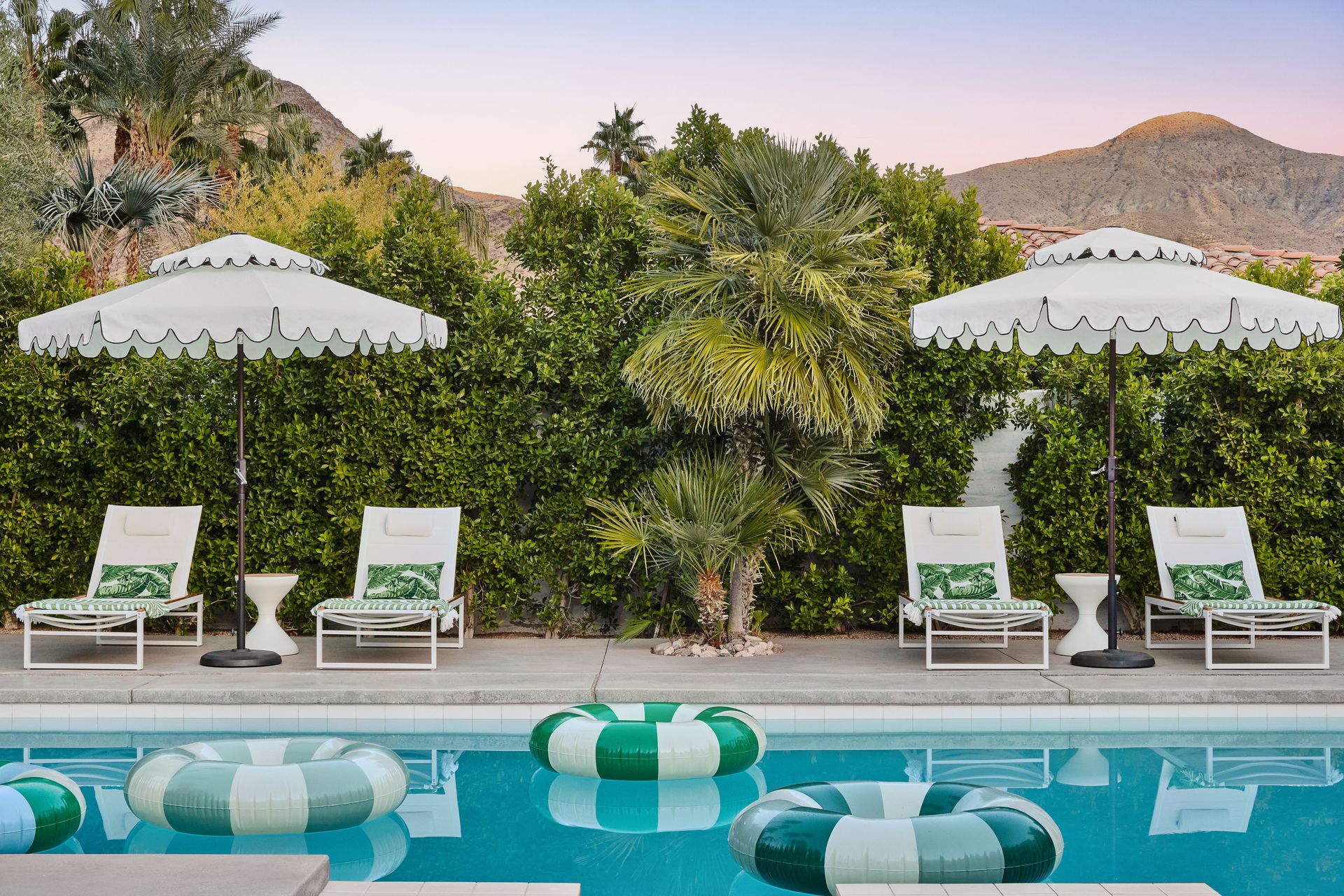 Poolside scene with lounge chairs, parasols, and floaties, backed by lush greenery and mountains.