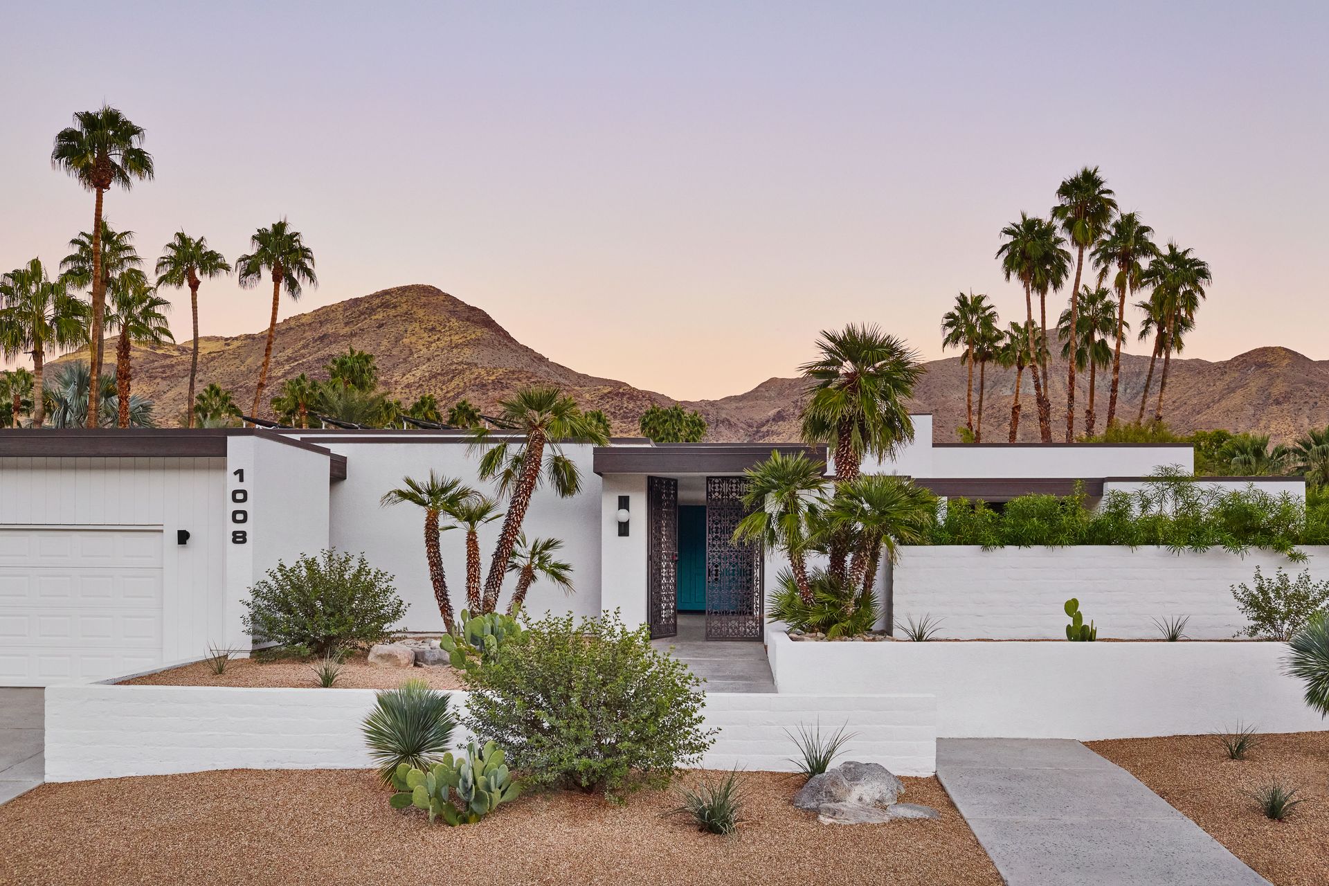 Mid-century modern house with palm trees, white walls, and mountain backdrop at dusk.