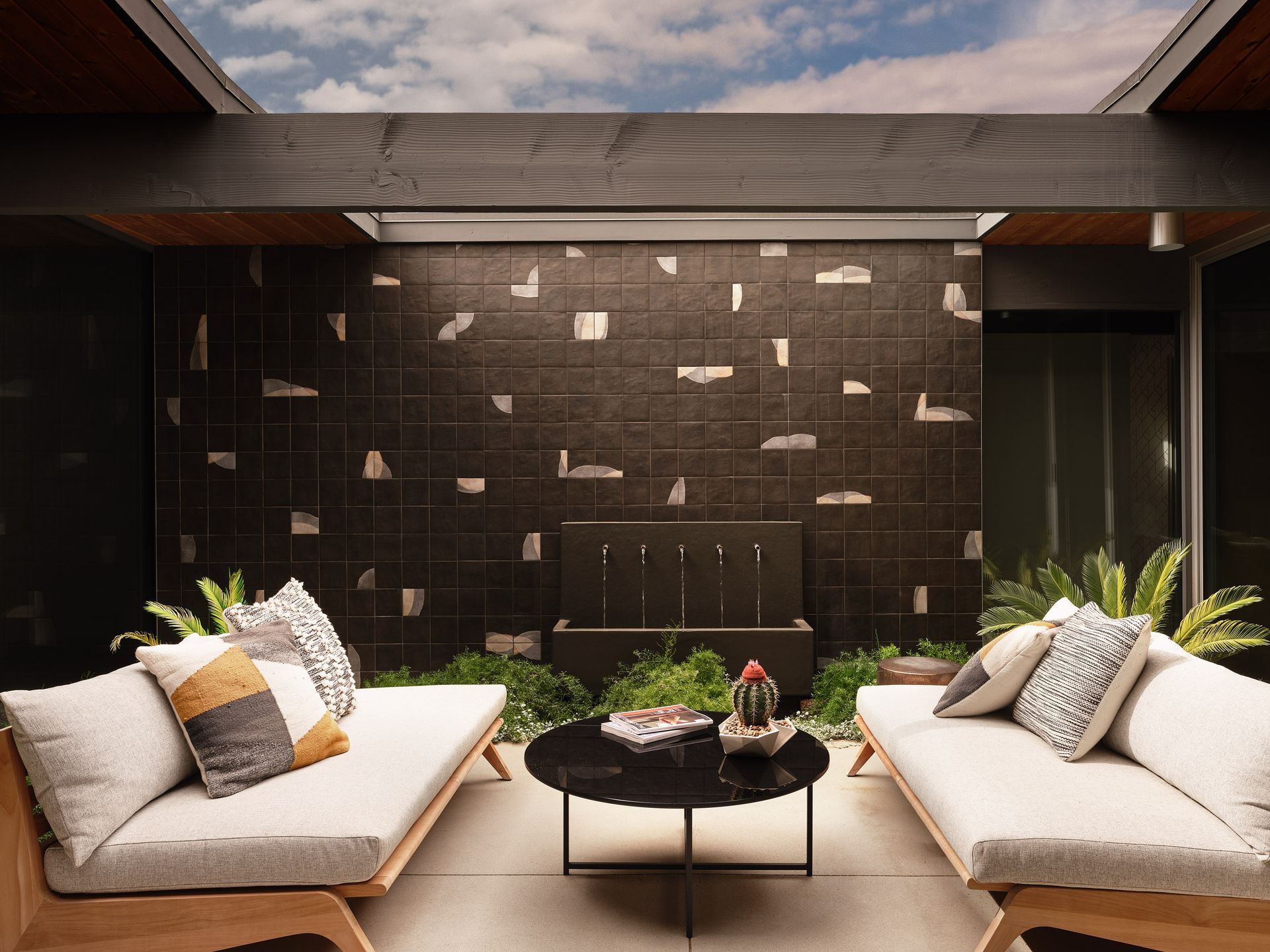 Outdoor patio with seating, dark tile wall, water feature, skylight, and greenery.