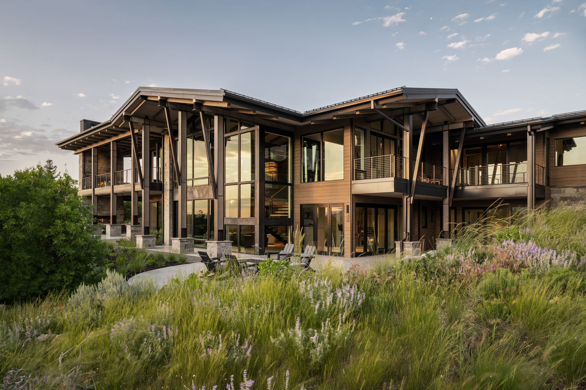 Large, modern house with glass walls and wooden beams, surrounded by tall grasses and flowers.