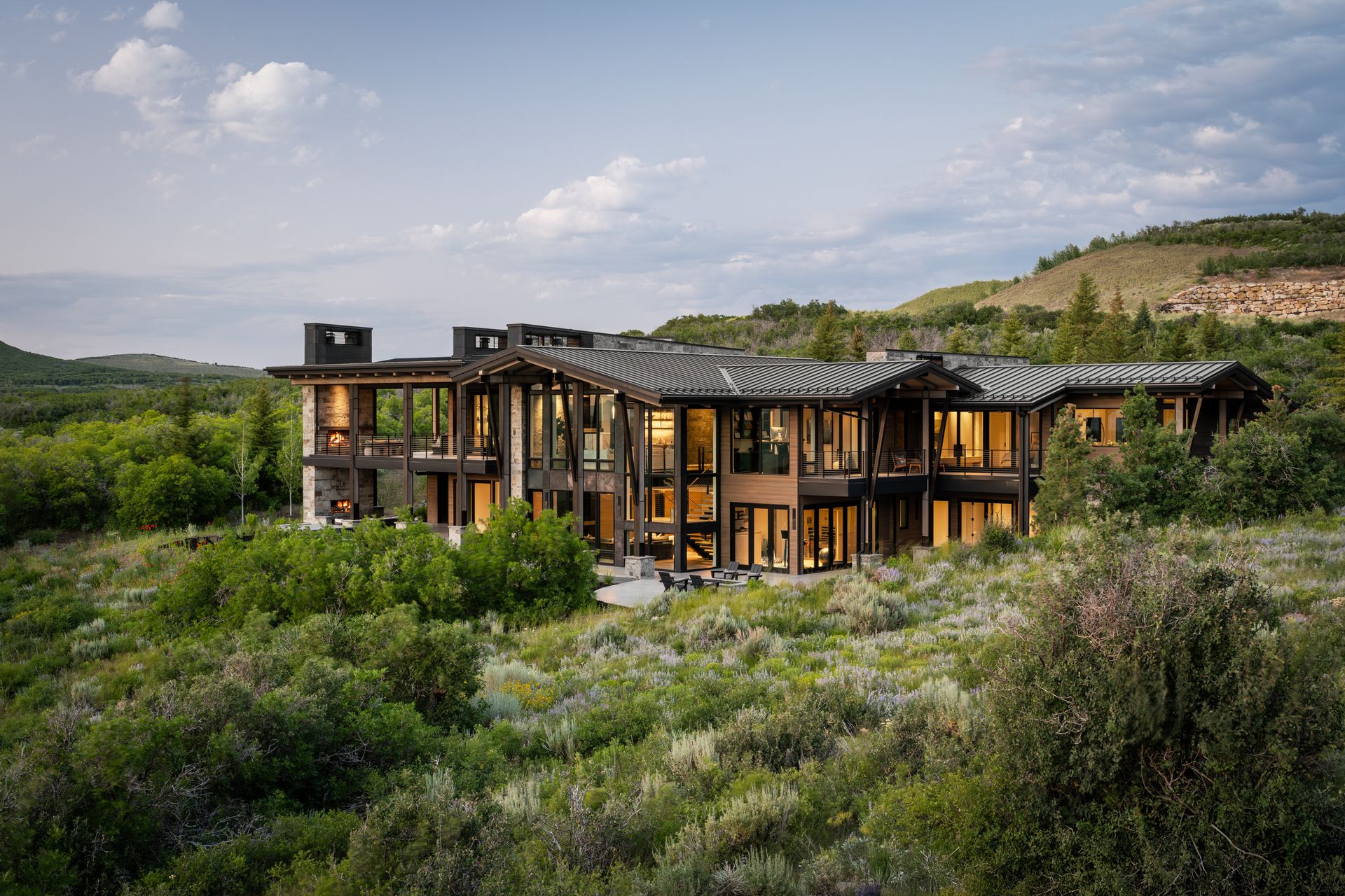 Large modern home nestled in a grassy, hillside landscape with dark wood and many windows.