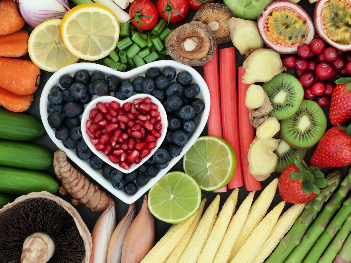 A heart shaped bowl of blueberries is surrounded by a variety of fruits and vegetables.