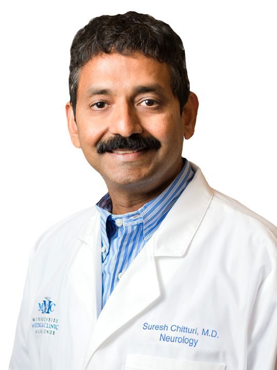 A man with a mustache is wearing a white lab coat and smiling.