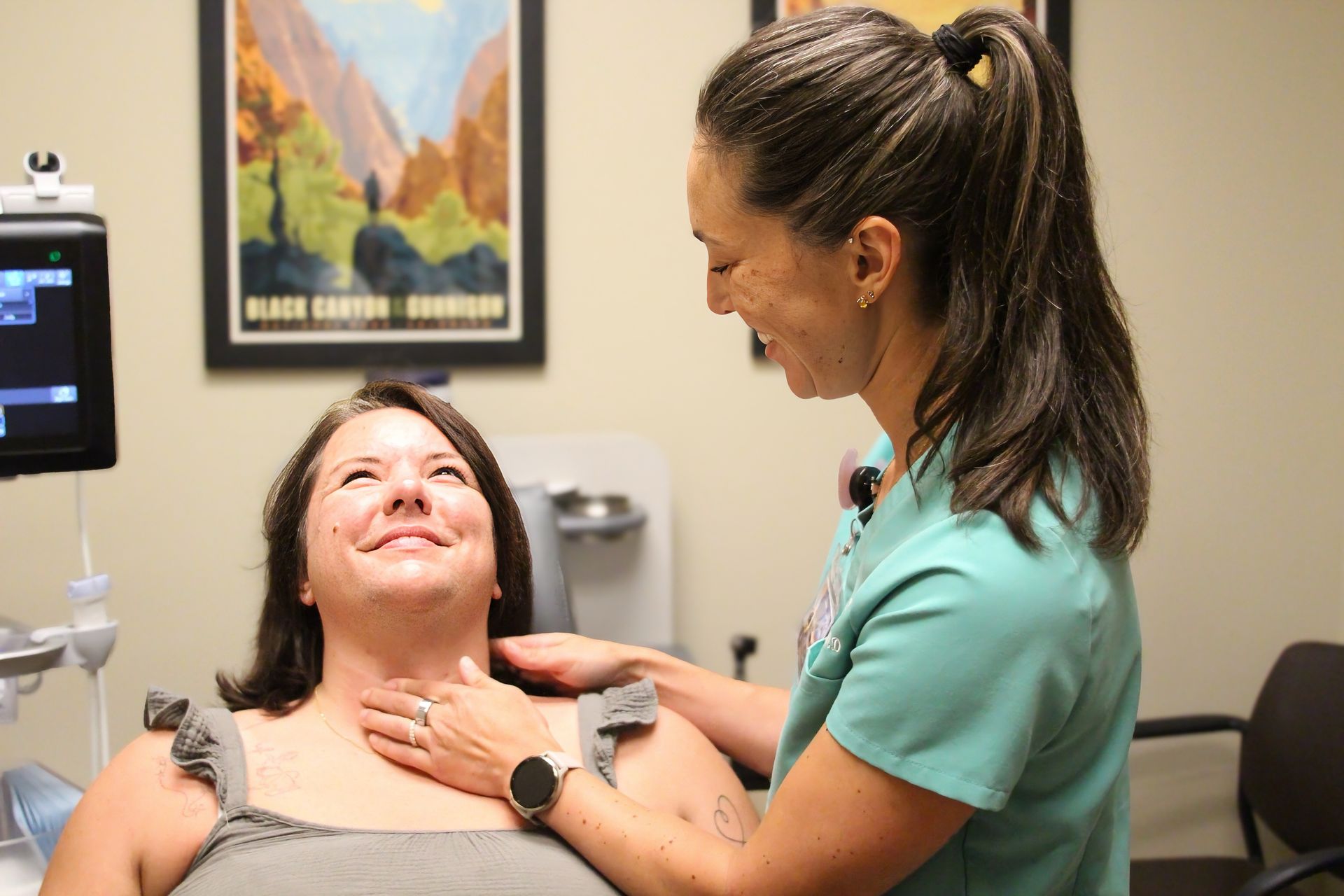 A smiling medical professional examines a patient's neck in an exam room, looking happy.