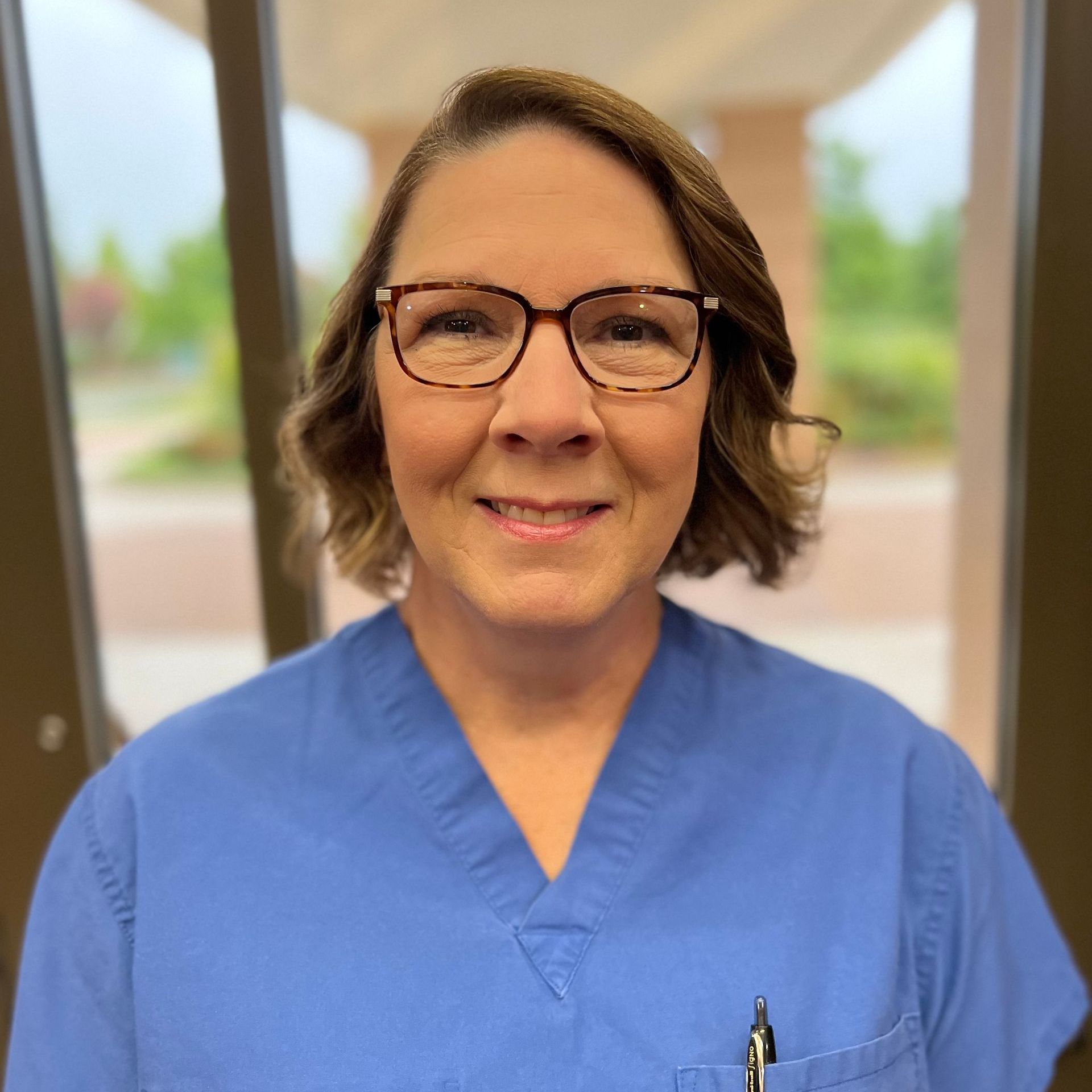 A woman wearing glasses and a blue scrub is smiling for the camera.