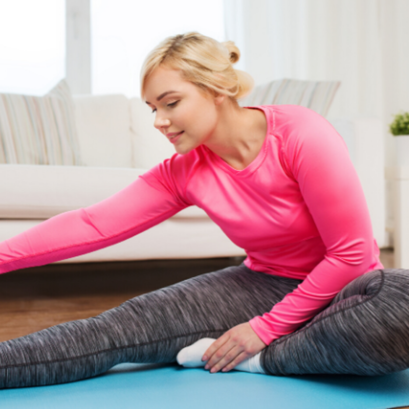 A woman in a pink shirt is stretching her legs on a yoga mat.