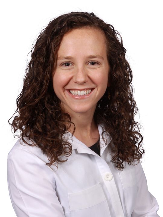 A woman with curly hair is wearing a white lab coat and smiling