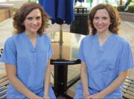 Two women in scrubs are sitting at a table under an umbrella.