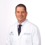 A man in a white lab coat and tie is standing in front of a white background.