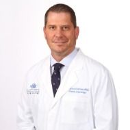 A man in a white coat and tie is standing in front of a white background.