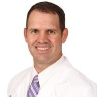 A man in a white coat and purple tie is smiling for the camera.
