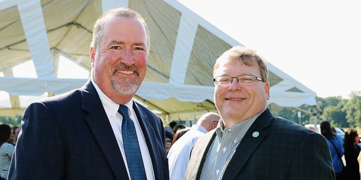 Two men in suits and ties are standing next to each other in front of a tent.