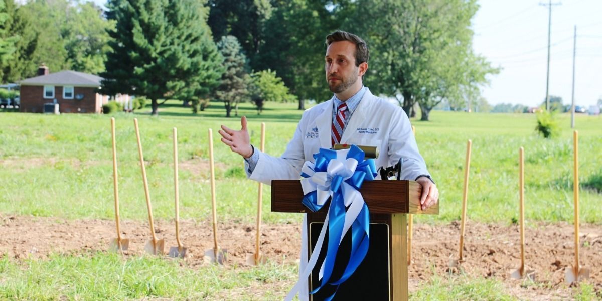 A man in a white coat and tie is standing at a podium in a field.