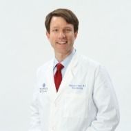 A man in a white lab coat and red tie is smiling.