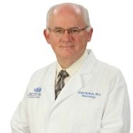 A man in a white lab coat and tie is standing in front of a white background.