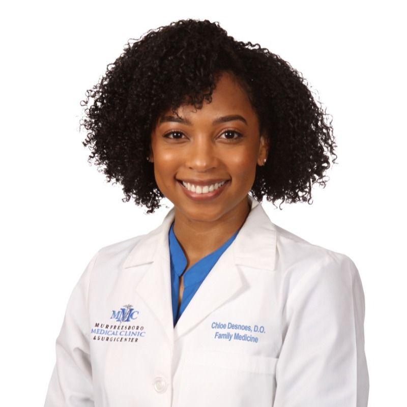 A woman wearing a white lab coat and blue scrubs is smiling