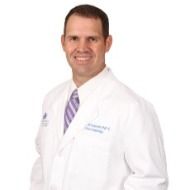 A man in a white lab coat and purple tie is smiling.