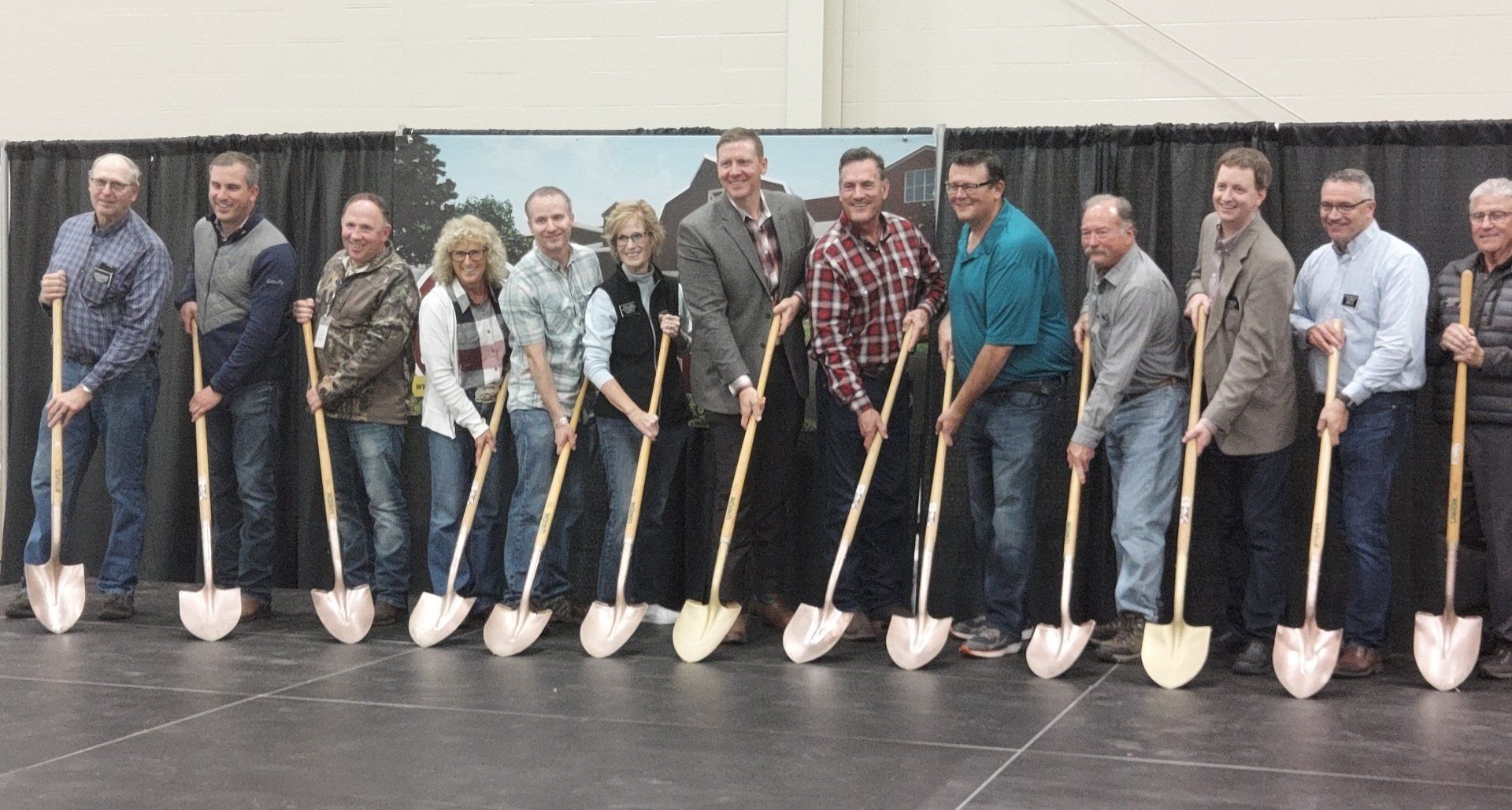 Group of people holding shovels, poised to break ground at an event. Stage setting.