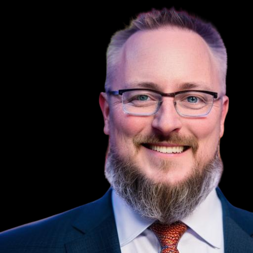 A person with a beard and glasses, smiling, wearing a blue suit, white shirt, and patterned tie against a black backdrop.