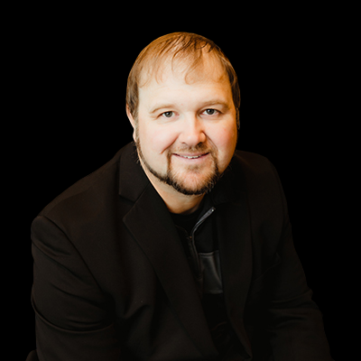 A person with short hair and facial hair, wearing a black blazer over a dark shirt against a black background.
