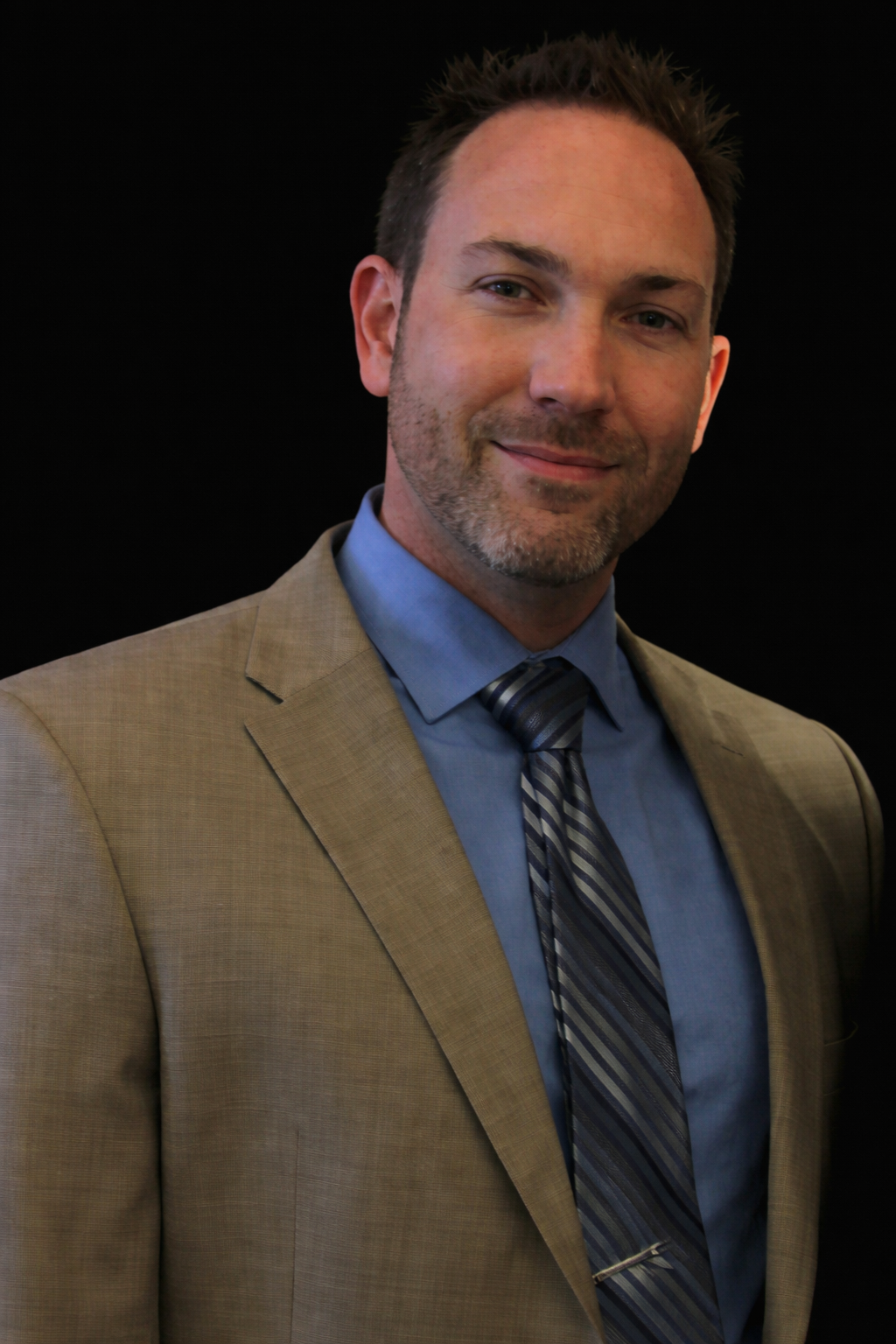 A person wearing a tan suit, blue shirt, and patterned tie against a black background.