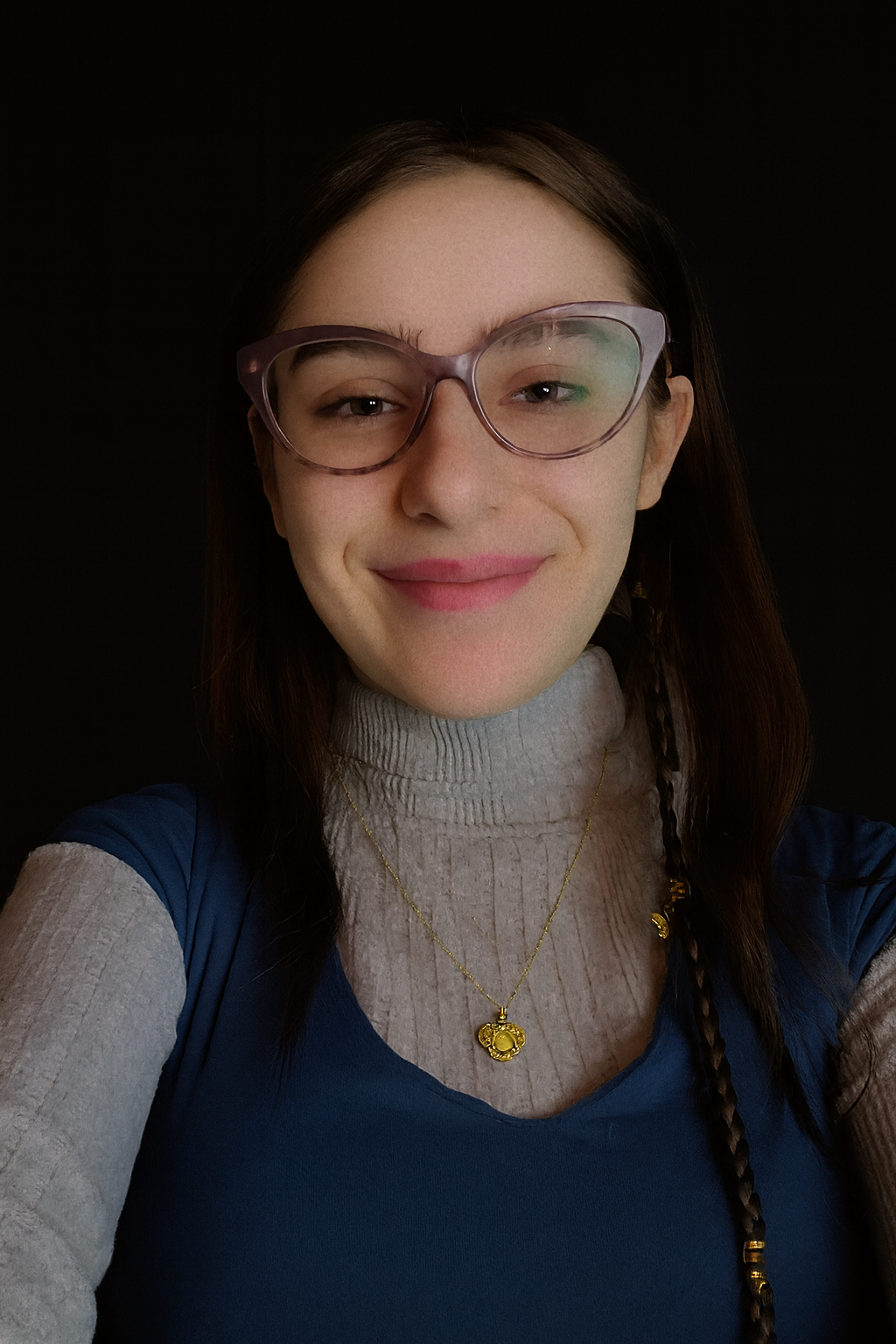A smiling person with long dark hair and cat-eye glasses wears a grey sweater and blue top against a black background.