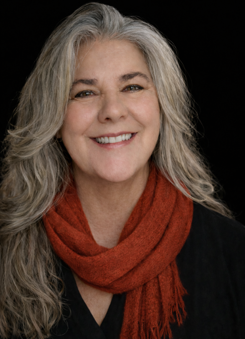 A person with long, gray hair and a rust-colored scarf smiles warmly in front of a blurred vintage typewriter background.